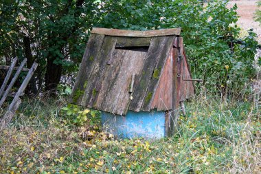 Old wooden water well with moss and peeling blue paint on the base stands forgotten amidst overgrown grass and foliage in a tranquil countryside landscape, evoking a sense of history and decay.