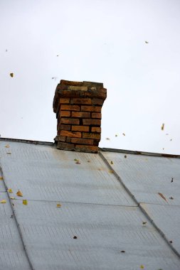 Eski mimarinin cazibesini ve sonbahar mevsiminin güzelliğini gözler önüne seren, yıpranmış metal bir çatının üzerinde, bulutlu gökyüzünün zeminine düşen sonbahar yapraklarıyla benekli, taşralı tuğla baca..