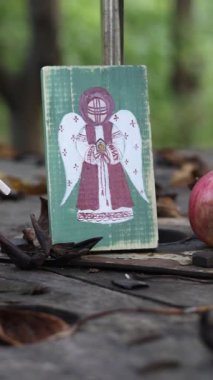 Kırsal ahşap bir masa üzerinde el boyaması bir melek figürü, küçük kırmızı bir ev, ve olgun bir elma, düşmüş yaprakların arasında, sakin, sonbahar havası yaratan, basit yaşam ve el sanatlarını kutlayan, mevsimsel dekor, kırsal cazibe.