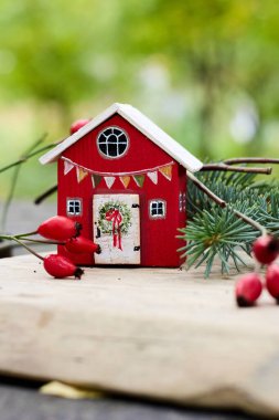 Beyaz çatı ve şenlikli çelenkle süslenmiş, canlı kırmızı böğürtlenler ve yemyeşil meyvelerle tamamlanmış kırmızı bir evin yer aldığı büyüleyici bir minyatür bayram sahnesi samimi ve şenlikli bir ortam yaratır..