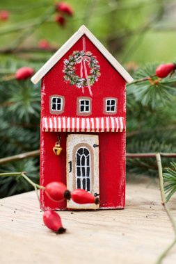 Şenlikli Durgun Hayat: Kırmızı Ahşap Oyuncak Evi 'nin Hoş Yakın Çekimi Noel süsleriyle süslenmiş, Kırsal Ahşap bir masa üstünde Rose Kalçaları tarafından tamamlanmış, Konforlu ve davetkar bir tatil atmosferi yaratan