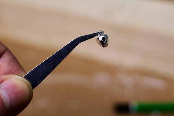 Детальный крупным планом захватывает руку деликатно держа крошечный серебряный колокольчик с помощью пинцета, подчеркивая его детальное мастерство на мягком, размытом фоне, подчеркивая точность.