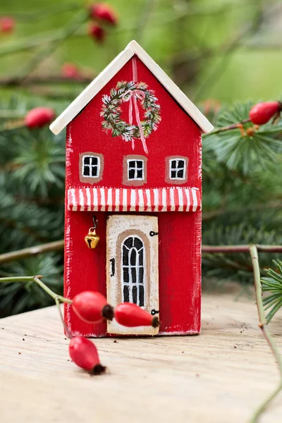 Şenlikli Durgun Hayat: Kırmızı Ahşap Oyuncak Evi 'nin Hoş Yakın Çekimi Noel süsleriyle süslenmiş, Kırsal Ahşap bir masa üstünde Rose Kalçaları tarafından tamamlanmış, Konforlu ve davetkar bir tatil atmosferi yaratan