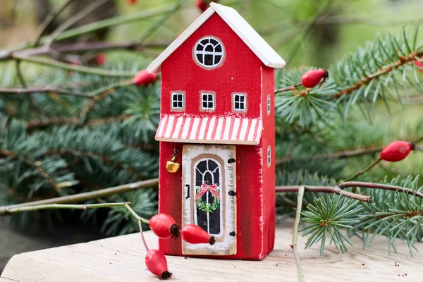 Şenlikli Noel sahnesinde el yapımı kırmızı ahşap bir ev, beyaz bir çatı ve güzel süslenmiş bir kapı, canlı köknar dalları ve canlı kırmızı gül kalçaları arasında yer alır..