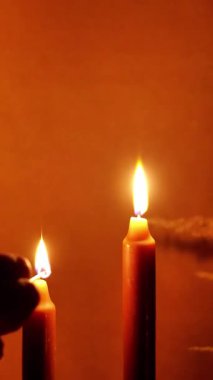 Aydınlatıcı sükunet: Süslü bir şamdanın içindeki üç kahverengi mumu zarifçe tutuşturan bir ele tanık olun, dingin ve davetkar bir ışıltı saçan sıcak turuncu bir arka plana.