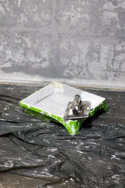 Kapalı alanda plastik bir çarşaf üzerinde dinlenen yeşil ve beyaz boya tepsisini yakına çekerken çekilmiş bir fotoğraf, sanatsal desenli duvar kaplamalı bir ev yenileme veya boyama projesine işaret ediyor. Tepsi korunmak için plastik bir çarşafa konur.