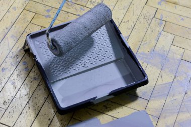 Yakın plan bir boya silindiri yakalıyor, gri boya dolu, bir boya tepsisi üzerinde duruyor, hepsi aynı renkte lekelerle işaretlenmiş ahşap parke zemine karşı, devam eden bir ev yenileme projesini gösteriyor..