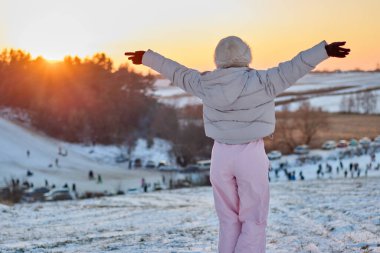 Kışlık paltolu ve şapkalı bir kadının, kolları açık kar kaplı bir tepenin üzerinde dikilip, gün batımında kar, kızak ve park halindeki arabalarla dolu kış manzarasını izlemesi..