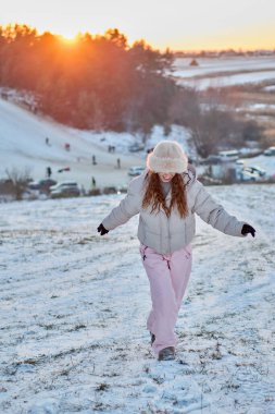 Kış kıyafetleri giymiş neşeli genç bir kadın gün batımında karla kaplı bir arazide yürüyor. Kış harikalar diyarının ortasında arabalarla ve uzaktan görülebilen insanlarla mutluluk saçıyor..