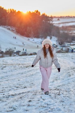 Gülümseyen kızıl saçlı kadın kış gününün tadını çıkarıyor. Kar tarlasında kürklü bir şapkayla, ceketiyle ve pembe kar pantolonuyla yürüyor. Altın saat boyunca, arkasında günbatımı..