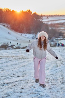 Kış kıyafetleri içinde kıpkırmızı saçları olan mutlu ve çekici genç bir kadın, neşeli bir ifadeyle kameraya bakarak, tepede koşarken gün batımının tadını çıkarıyor.