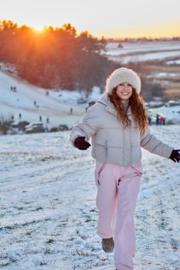 Sıcak kış kıyafetleri giymiş, kürk şapkası ve pembe kar pantolonu da dahil mutlu bir genç kadın, gün batımında karla kaplı bir tepeyi zevkle keşfediyor ve kış manzarasına eğlence ve özgürlük getiriyor..