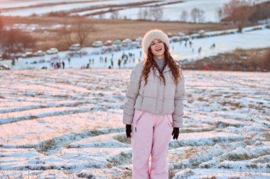 Güzel kız kışın gülümsüyor ve poz veriyor, kürk şapka ve kabarık bir ceket giyiyor, karla kaplı doğa aktivitelerinin tadını çıkarıyor eğlence ve mutlulukla..