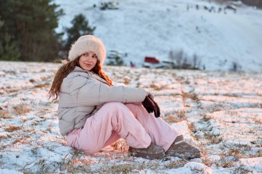 Kış kıyafetleri içinde mutlu bir genç kadın karla kaplı bir tarlada oturur, güneşli güzel bir günün tadını çıkarır ve etrafındaki çarpıcı kış manzarasının tadını çıkarır, rahat ve mutlu hisseder..
