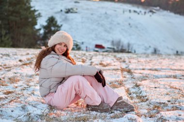 Gülümseyen genç bir kadın kış günbatımının tadını çıkarırken kar kaplı bir tepede kürk şapka, kabarık bir ceket ve kış pantolonu giyerek mükemmel bir kış manzarası yaratıyor..