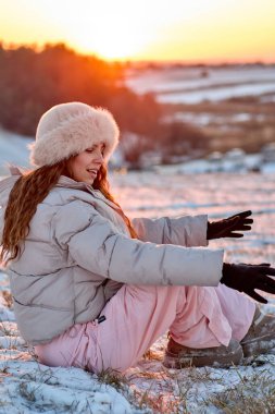 Kış manzarasında kürk şapkalı ve kabarık ceketli bir kadın, açık kollarıyla karda oturmuş, güneşin sıcacık renklerinin tadını çıkarıyor, kış mutluluğunun özünü yakalıyor..
