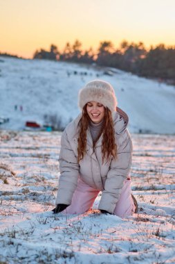 Rahat bir kış elbisesi içindeki neşeli genç bir kadın karda diz çöker, gülüşü gün batımında sakin bir kar manzarasının zemininde ışıl ışıl parlar, kışın mutluluğunu ve doğal güzelliğini yakalar..