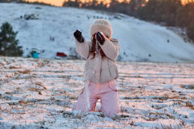 Kış şapkası ve paltosu giymiş mutlu bir çocuk, parlak ve güneşli bir kış gününde dizlerinin üstüne çöküp havaya kar atıyor. Gündüz vakti arka planda karla kaplı tepeler var..