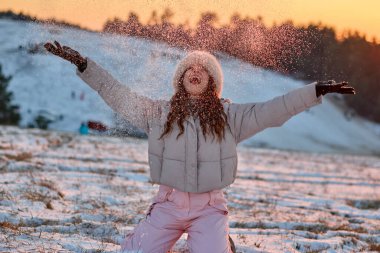 Kışın giyinmiş neşeli genç bir kadın, karlı bir tarlada, gün batımında diz çöker. Kollarını açıp karları kucaklayarak kışın mutluluğunu yakalar..