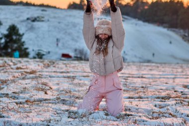 Kürk şapkalı neşeli genç kadın kışın gün batımında karla kaplı bir çayırda diz çökerken havaya kar fırlatıyor, doğada kış tatillerini kutluyor..