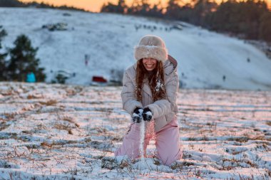Mutlu ve gülümseyen genç bir kadın, kar kaplı bir parkta güneşli bir günün tadını çıkarırken kürk şapka, kışlık ceket ve pembe kayak pantolonu giyerek kışın eğleniyor..