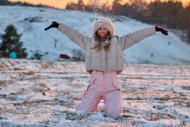 Güzel bir kız kış harikalar diyarının güzelliğinin tadını çıkarıyor, karda neşeyle diz çökmüş, elleri karla dolu, güneş batarken ışıl ışıl gülümserken..