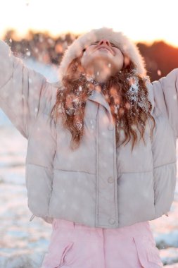 Mutlu genç bir kadın kar yağışından zevk alır, yüzü kışın gün batımında, rahat pembe bir kabarık ceket ve tüylü bir şapka giyerek oynarken keyifle yukarı doğru eğilir..