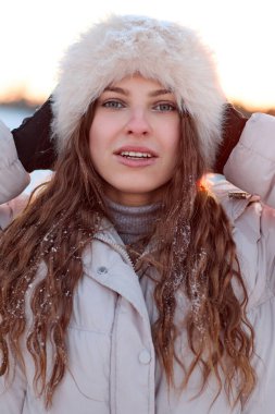 Kışlık giysiler giymiş, kar kaplı beyaz tüylü bir şapka takmış güneşli bir kış gününde kameraya gülümseyen ve eldivenli elleriyle başını tutan çekici bir kadın..
