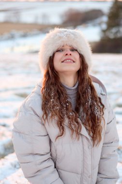 Kış kıyafetleri giymiş çekici bir kadın, parlak bir gülümseme ile kış gününün tadını çıkarır, kar tozunu saçlarına ve ceketine sergiler, karlı bir arka planda, bulanık bir şekilde dikilirken..