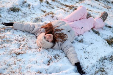 Kış şapkası ve şık pembe bir kayak elbisesi giyen hoş genç bir kadın, neşe içinde ışıldayan karda bir kar meleği yaratıyor, yüzü mutlulukla parlıyor, kışın ve özgürlüğün özünü yakalıyor..