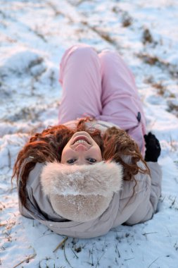 Karda sırtüstü uzanmış, kış gününün keyfini çıkartan neşeli genç bir kadın. Kürk şapkası ve güzel bir gülümsemeyle, rahat bir mevsim portresi yaratıyor..