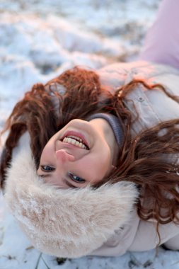 Güzel, gülümseyen mavi gözlü genç bir kadın kış günü kürk şapka ve kışlık palto giyerek sırtüstü uzanıyor. Yüzü yakın, göz teması kuruyor, mutlu ve neşeli..