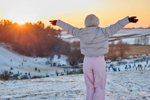 Kışlık paltolu ve şapkalı bir kadının, kolları açık kar kaplı bir tepenin üzerinde dikilip, gün batımında kar, kızak ve park halindeki arabalarla dolu kış manzarasını izlemesi..