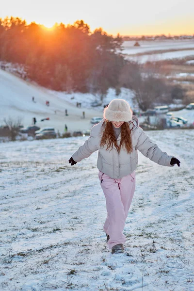 Kış kıyafetleri giymiş neşeli genç bir kadın gün batımında karla kaplı bir arazide yürüyor. Kış harikalar diyarının ortasında arabalarla ve uzaktan görülebilen insanlarla mutluluk saçıyor..