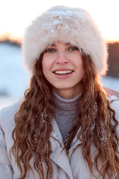 Çekici genç bir kadın kış günbatımını kürk şapkası ve kalın bir ceket giyerek seviyor. Gülümseyen yüzü, kar tanelerinin açık havada saçlarını süslemesi gibi neşe saçıyor..