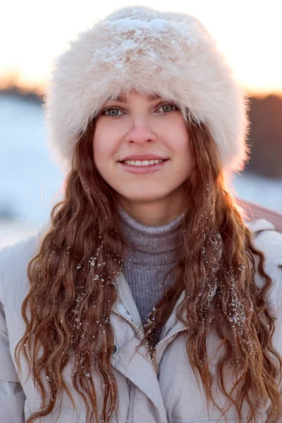 Güzel, genç bir kadın kışın dışarıda poz veriyor. Kürk şapkası ve kalın bir ceket giyiyor. Uzun, kıvırcık saçları narin kar taneleriyle süslenmiş..