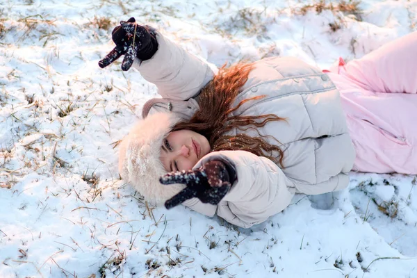 Kış ceketi ve kürk şapkalı gülümseyen genç kadın karda uzanıyor, eldivenli elleriyle barış işareti yapıyor, kış gününün tadını çıkarıyor ve tabiatında iyi bir ruh hali var..