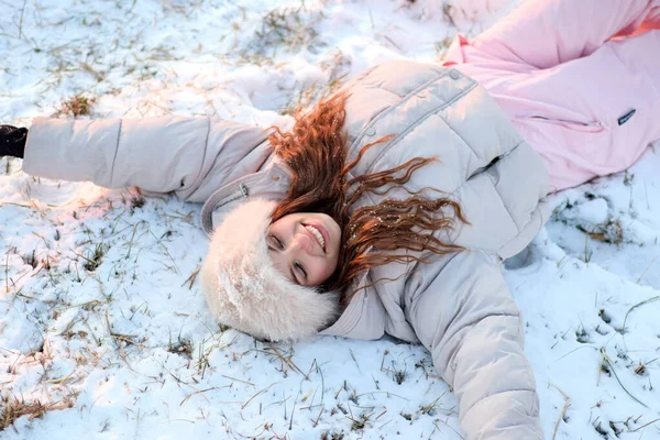 Dalgalı kahverengi saçlı ve şık tüylü şapkalı neşeli genç bir kadın, karlı arazide yatarken mutlu bir şekilde gülümsüyor, şık bej renkli bir kabarık ceket ve pembe bir kayak pantolonu giyiyor, keyifli bir kış anının özünü yakalıyor..