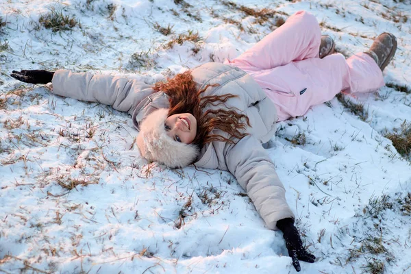 Güzel ve gülümseyen genç bir kadın kışlık şapka, sıcak bir ceket ve pembe bir kayak pantolonu giyer, karda uzanır, mutlu bir kış günü geçirir ve neşeyle kameraya bakar..