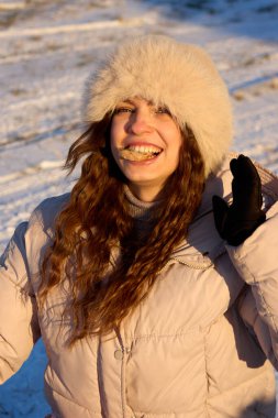 Kış kıyafetleri içinde kürk şapka ve kabarık palto giymiş, kumral kıvırcık saçlı neşeli genç bir kadın kar kaplı bir tarlada ekmeği yerken mutlu bir şekilde göz teması kuruyor..