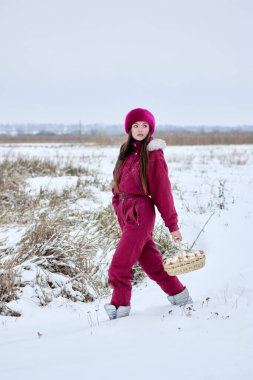 Canlı pembe bir kar elbisesi ve eşleşen bir kürk şapka giymiş genç bir kadın, dikkatle kar kaplı bir alanda yürüyor, sepet dolusu taze toplanmış beyaz mantarlar taşıyor..