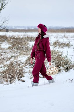Kestane rengi tulum giymiş ve kürk şapkalı genç bir kadın bir kış günü karlı bir tarlada yürüyor, bir sepet dolusu taze toplanmış yumurtayı taşıyor, dingin kış manzarasının tadını çıkarıyor..