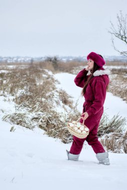 Zarif bir şekilde kar kaplı bir arazide şık bir kestane rengi kar elbisesi ve lüks bir kürk şapkayla yürüyen genç bir kadının büyüleyici görüntüsü. Süslü yumurtalarla dolu büyüleyici bir sepet taşıyor.