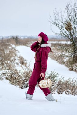 Huzurlu bir kış sahnesinde, kırmızı kar elbisesi ve zarif pembe kürk şapkalı genç bir kadın beyaz yumurtalarla dolu bir sepeti taşırken zarifçe karla kaplı bir arazide yürüyor..
