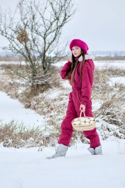 Canlı marsala kıyafetli genç bir kadının büyüleyici görüntüsü. Kar kaplı bir arazide zarif bir şekilde yürüyor. Beyaz şekerlemelerle dolu büyüleyici bir sepet taşıyor..