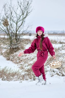 Canlı pembe bir kar elbisesi içinde büyüleyici bir kadın kış harikalar diyarında geziniyor, beyaz küresel nesnelerle dolu bir sepeti sallıyor, kürk şapkası ve botlarıyla tamamlanmış, neşeyle karlı manzarayı kucaklıyor..
