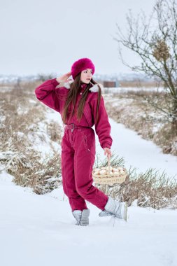 Canlı kırmızı bir kayak elbisesi, şık bir kürk şapkası ve zarif gümüş çizmeleri olan çekici genç bir kadın zarif bir sepetle güzel beyaz şekerlemelerle dolu bir sepet taşıyor karlı bir manzarada kışın cazibesinin özünü yakalıyor..