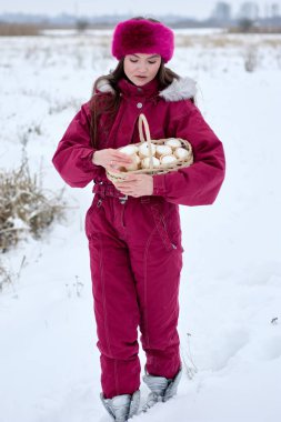 Canlı mor bir kar elbisesi içindeki genç kadın, uygun bir kürk bandıyla tamamlanmış, nazikçe bir sepet dolusu çöreği karlı bir manzaranın ortasına yerleştirir, bir kış günü aşağıya bakar..