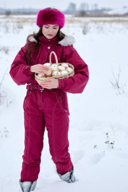Bordo bir kış tulumu giymiş genç bir kadın, elinde geleneksel paskalya çörekleriyle dolu bir sepet tutarak karda duruyor. Kış ormanı zeminine kurulmuş, sakin bir Paskalya kutlamasını simgeliyor..