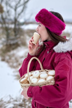 Tüylü kapüşonlu canlı kırmızı kış ceketi giymiş çekici genç bir kadın, şık bir kürk bandıyla süslenmiş, karlı bir arka planda kremalı beyaz bir dondurma külahının tadını çıkarıyor..